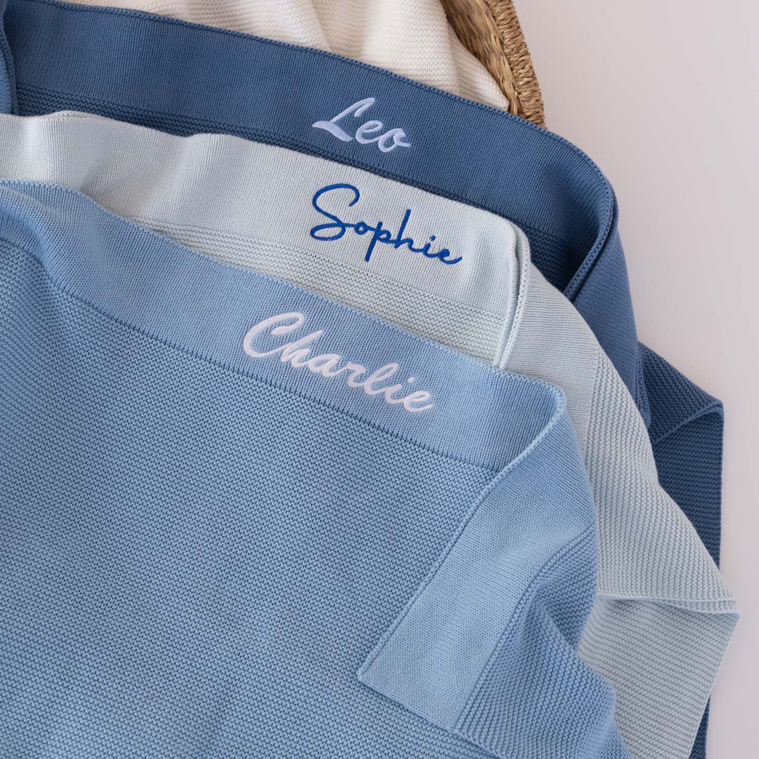 Three personalised knitted baby blankets in dark blue, light grey blue and sky blue showing shade comparison, each with embroidered script names in contrasting thread colours.