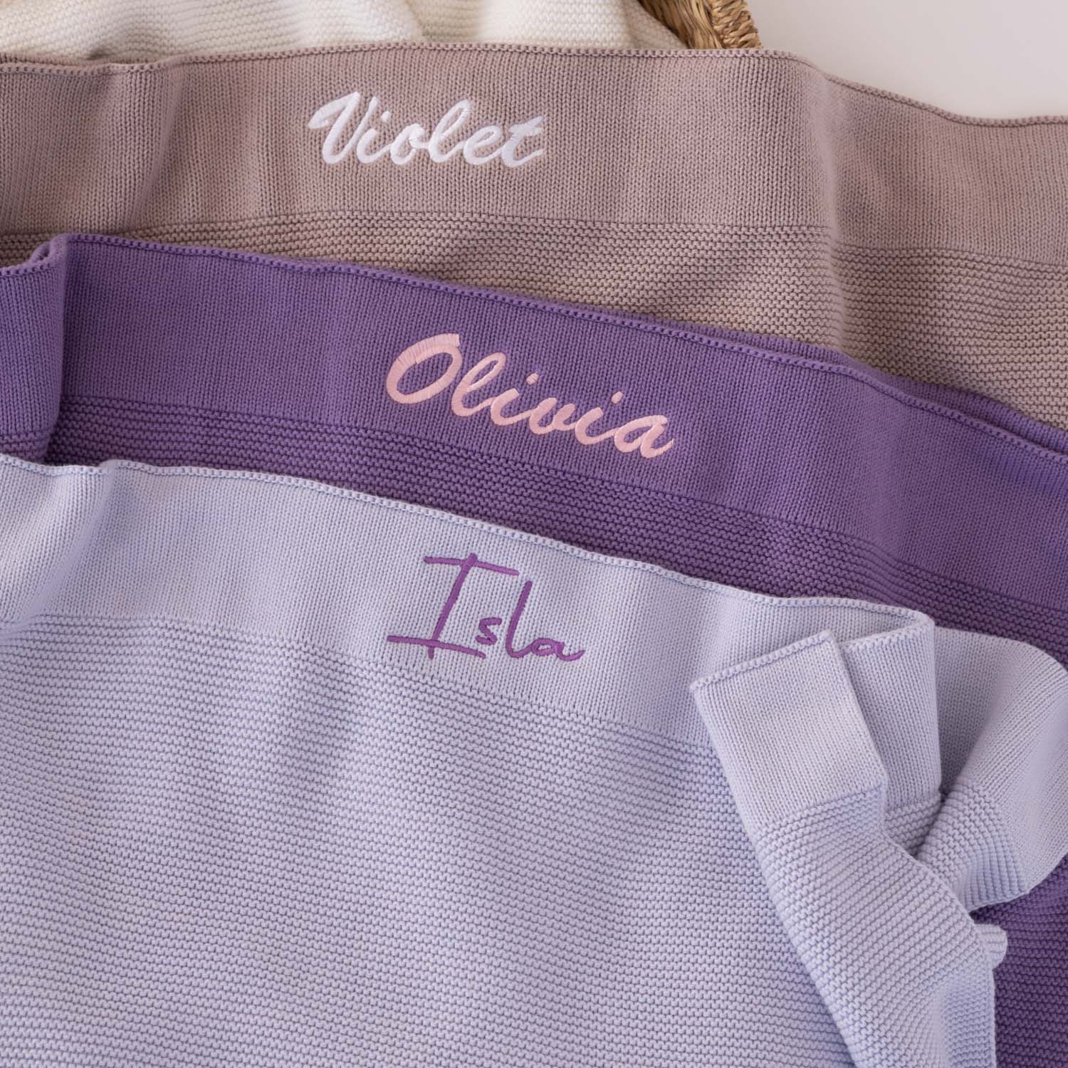 Three personalised knitted baby blankets stacked in lavender, deep purple and dusty lilac with embroidered script names in contrasting thread colours.