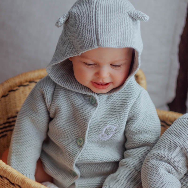 Smiling baby sitting in a wicker basket wearing a personalised grey green sage hooded knitted cardigan with bear ears embroidered with the initial G.