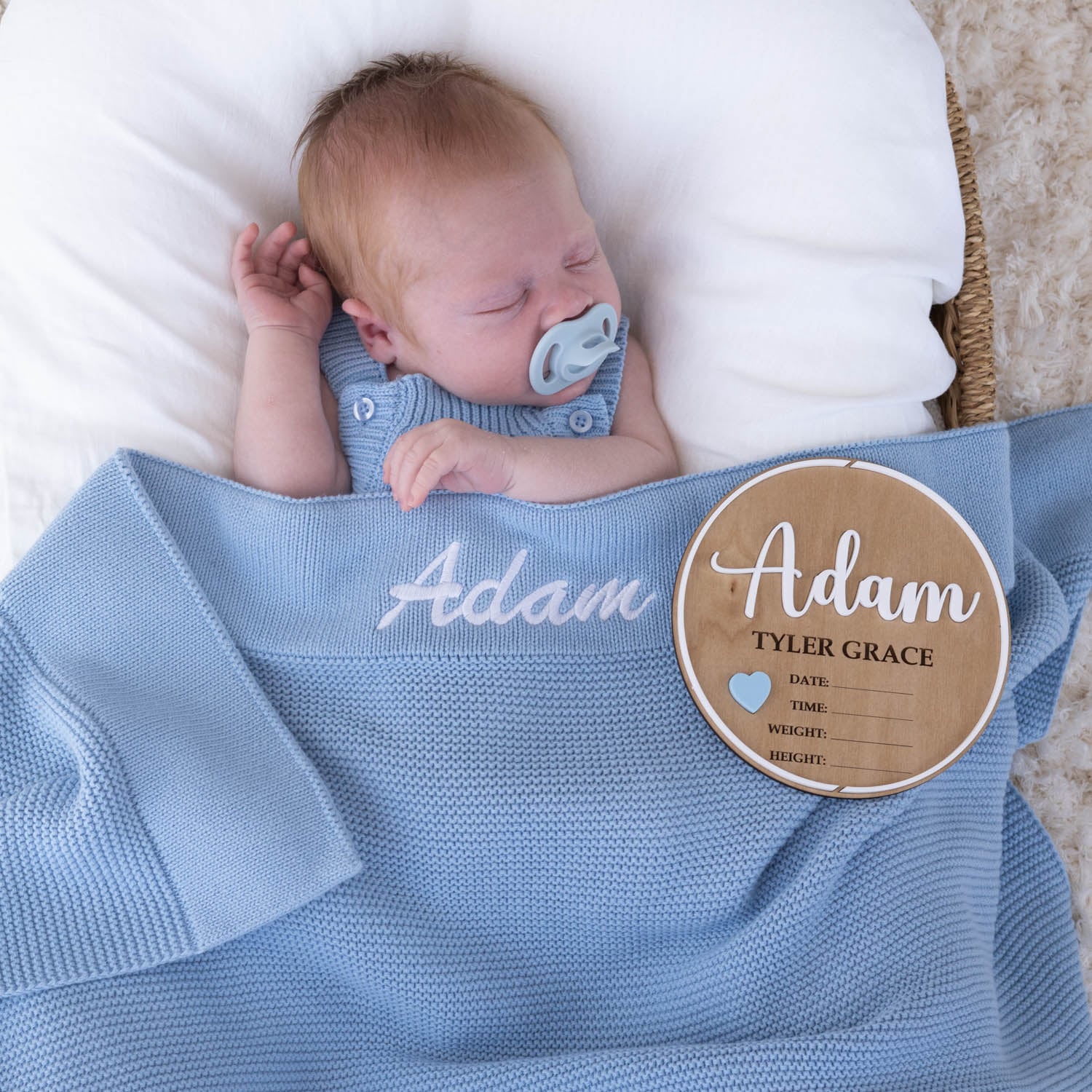 Personalised light blue knitted blanket with Adam embroidered in white on sleeping newborn beside wooden nursery plaque with Adam Tyler Grace and blue heart detail