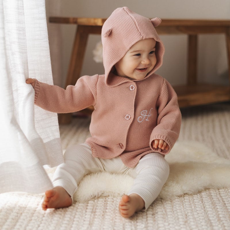 Smiling baby wearing a personalised blush pink hooded knitted cardigan with bear ears and embroidered H initial.