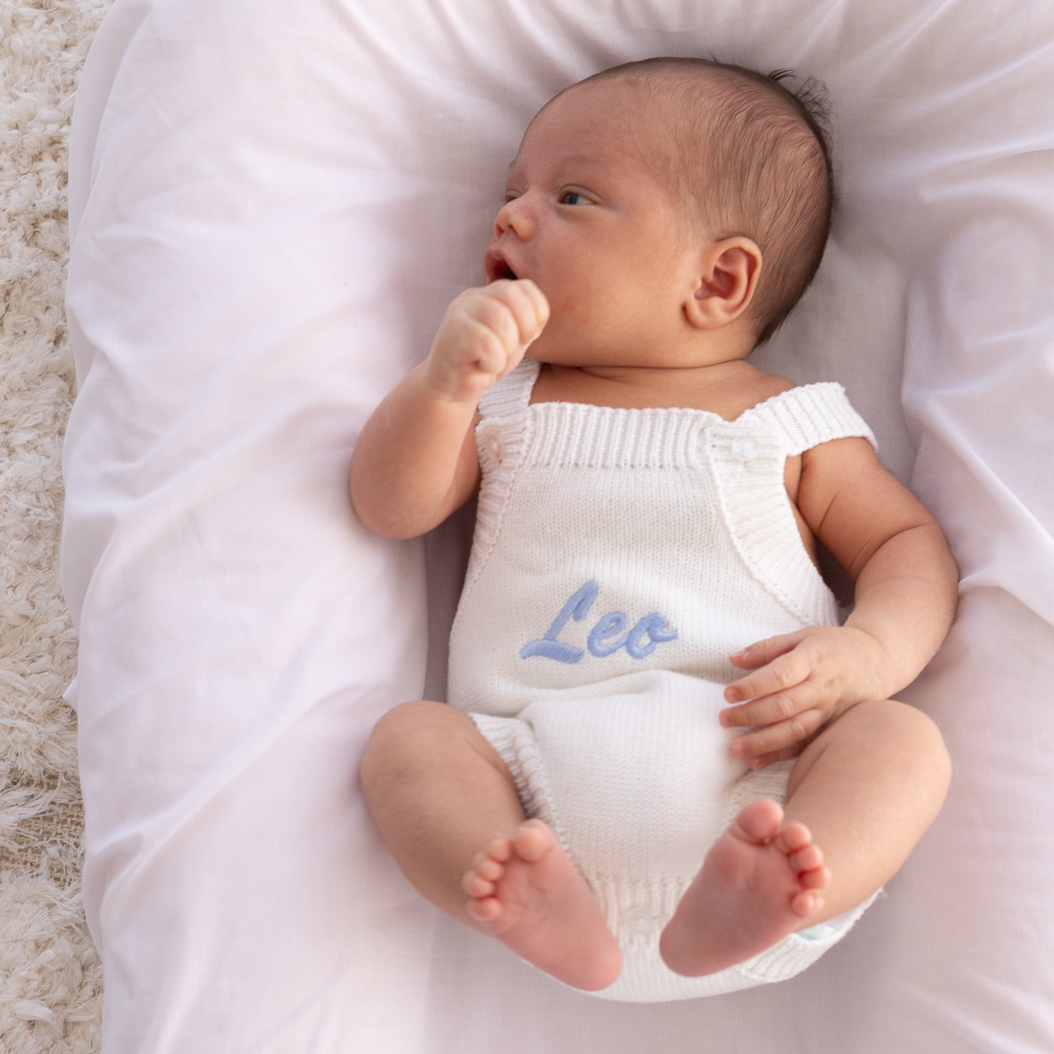 Personalised white knitted romper with Leo embroidered in blue on newborn lying on bassinet sheets